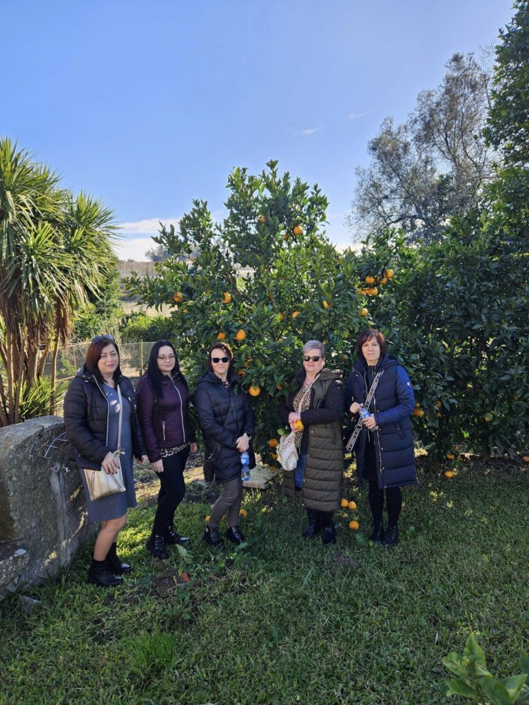Öt nőből álló csoport egy narancsfa előtt a kertben egy napsütéses napon. Kabátba és kabátba vannak öltözve, néhány táskában, egyben pedig műanyag palack. A háttérben buja növényzet és tiszta kék ég látható.