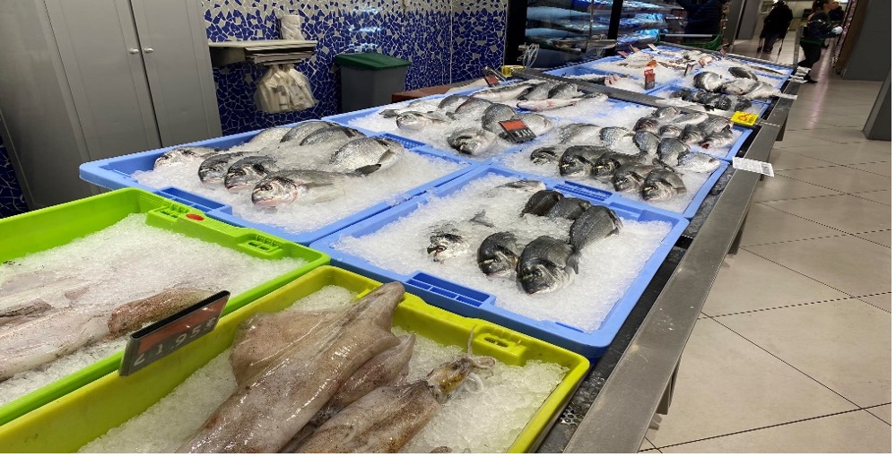Egy szupermarket halpultja, ahol különféle friss halak vannak jégen. A különböző típusú halakat kék és zöld tálcákba helyezzük. Néhány tálcán árcímkék láthatók. A háttérben több tengeri termék és egy böngésző személy látható.