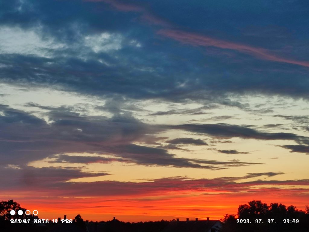 Élénk naplemente narancssárga, piros és kék árnyalatokkal festi az eget. A sötét felhők szétszóródnak, drámai hatást keltve. Alul a háztetők és a fák sziluettje látható. A képen látható szöveg a dátumot, "2023. 07. 07. 29:49" és a használt készüléket jelzi: "REDMI NOTE 10 PRO.