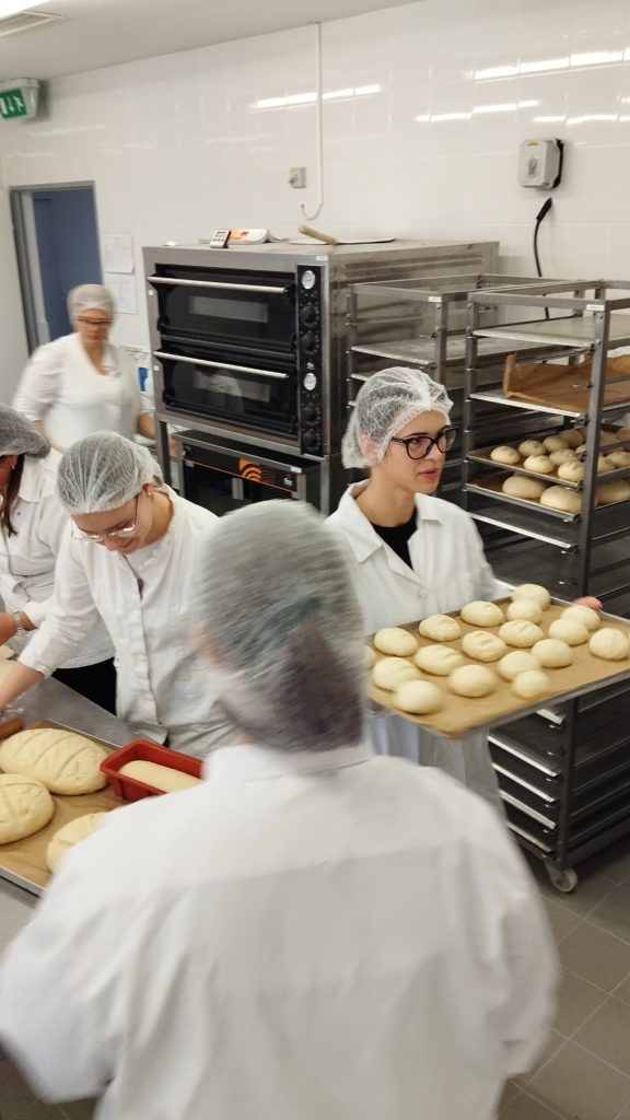Fehér egyenruhát és hajhálót viselő pékek egy csoportja dolgozik egy kereskedelmi konyhában. Az egyik pék a kezében tart egy tálcát sütetlen kenyérrel. A háttérben egy nagy sütő, valamint tésztával és pékáruval töltött rácsok láthatók.