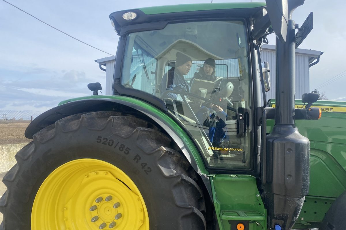 Két ember ül egy zöld traktor vezetőfülkéjében egy farmon. Mosolyognak és beszélgetnek. A traktor nagy, sárga kerekekkel rendelkezik, és egy épület közelében parkol, részben felhős égbolt alatt. A jelenet azt sugallja, hogy vagy vezetni tanulnak, vagy mezőgazdasági munkára készülnek.