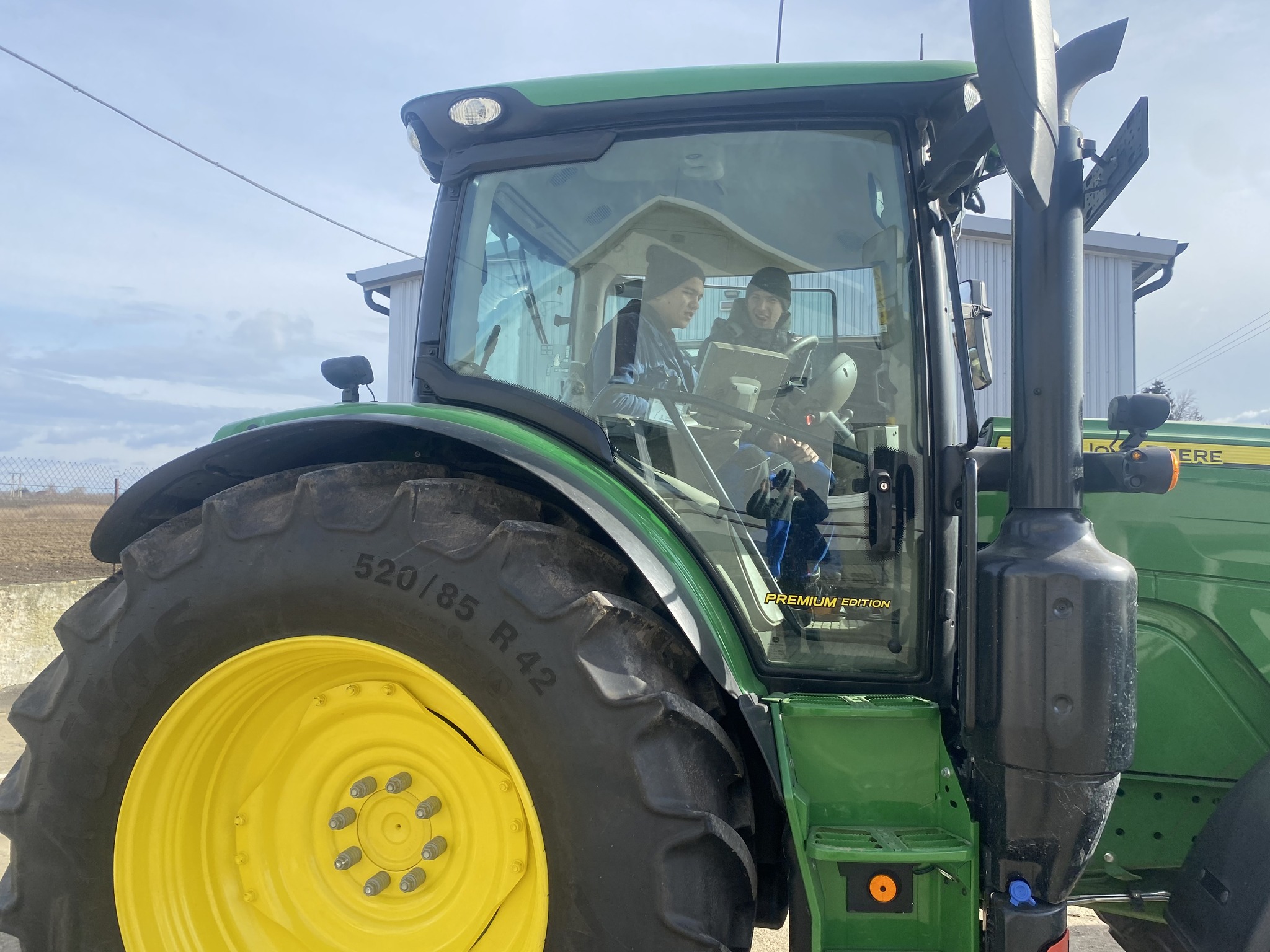 Két ember ül egy zöld traktor vezetőfülkéjében egy farmon. Mosolyognak és beszélgetnek. A traktor nagy, sárga kerekekkel rendelkezik, és egy épület közelében parkol, részben felhős égbolt alatt. A jelenet azt sugallja, hogy vagy vezetni tanulnak, vagy mezőgazdasági munkára készülnek.