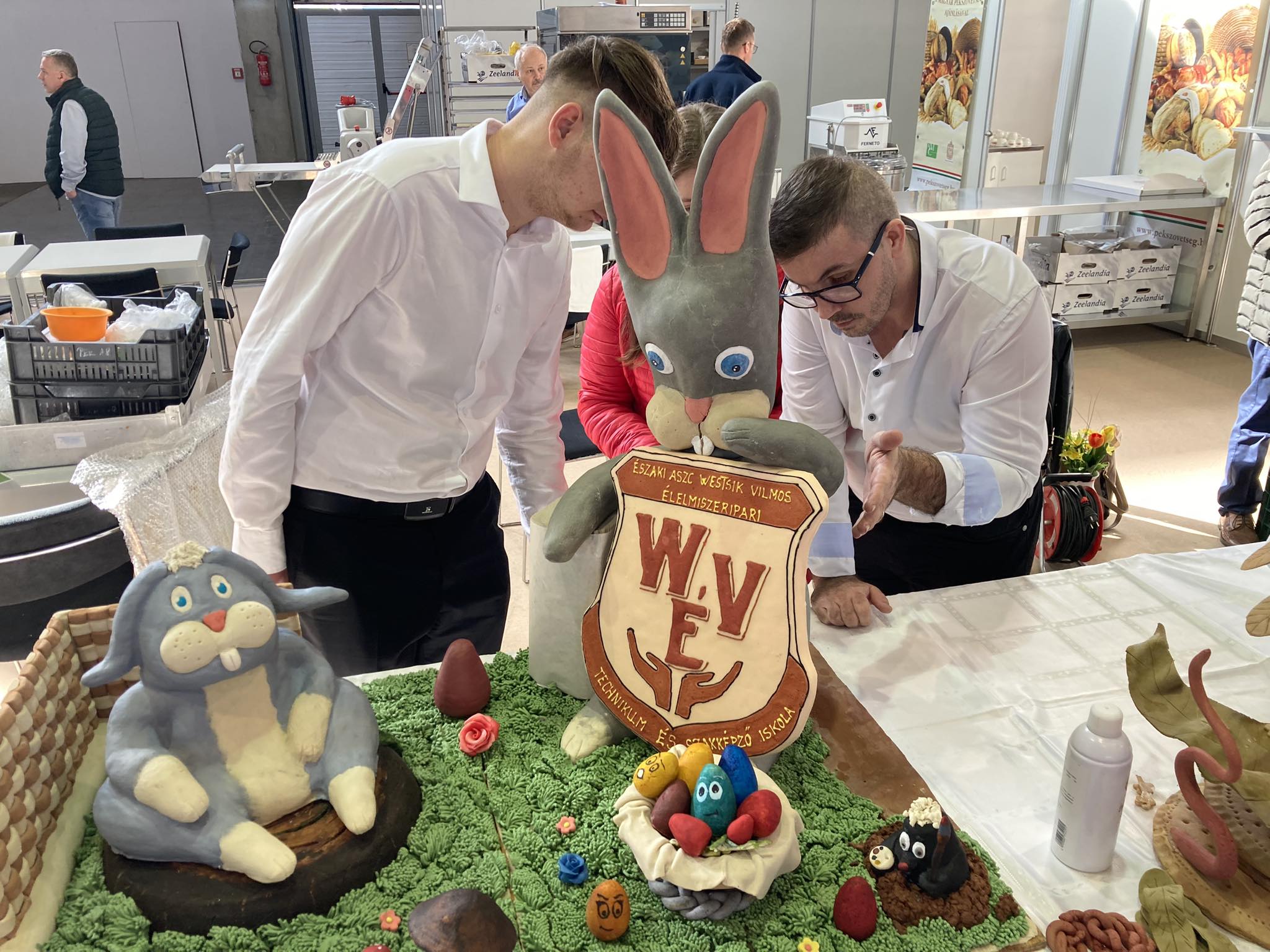 Három ember egy bonyolult tortakiállítást vizsgál egy rendezvényteremben. A tortán egy nagy nyúl látható, aki egy címert tart a "W.E.Y" kezdőbetűkkel. és kisebb fondant állatok füves alapon. A jelenet egy sütőversenyt vagy kirakat beállítást jelez.