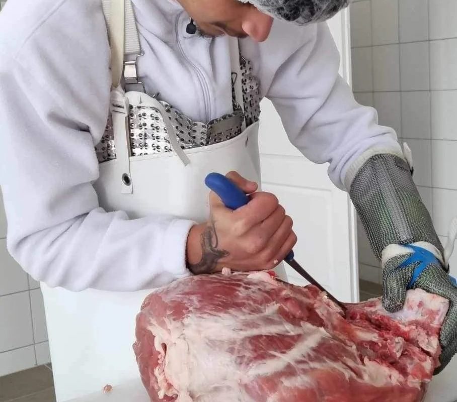 Egy fehér kabátot, védőkesztyűt és hajhálót viselő személy késsel vág le egy nagy darab húst. Úgy tűnik, hogy a helyszín egy tiszta, fehér csempézett hentes vagy konyha.