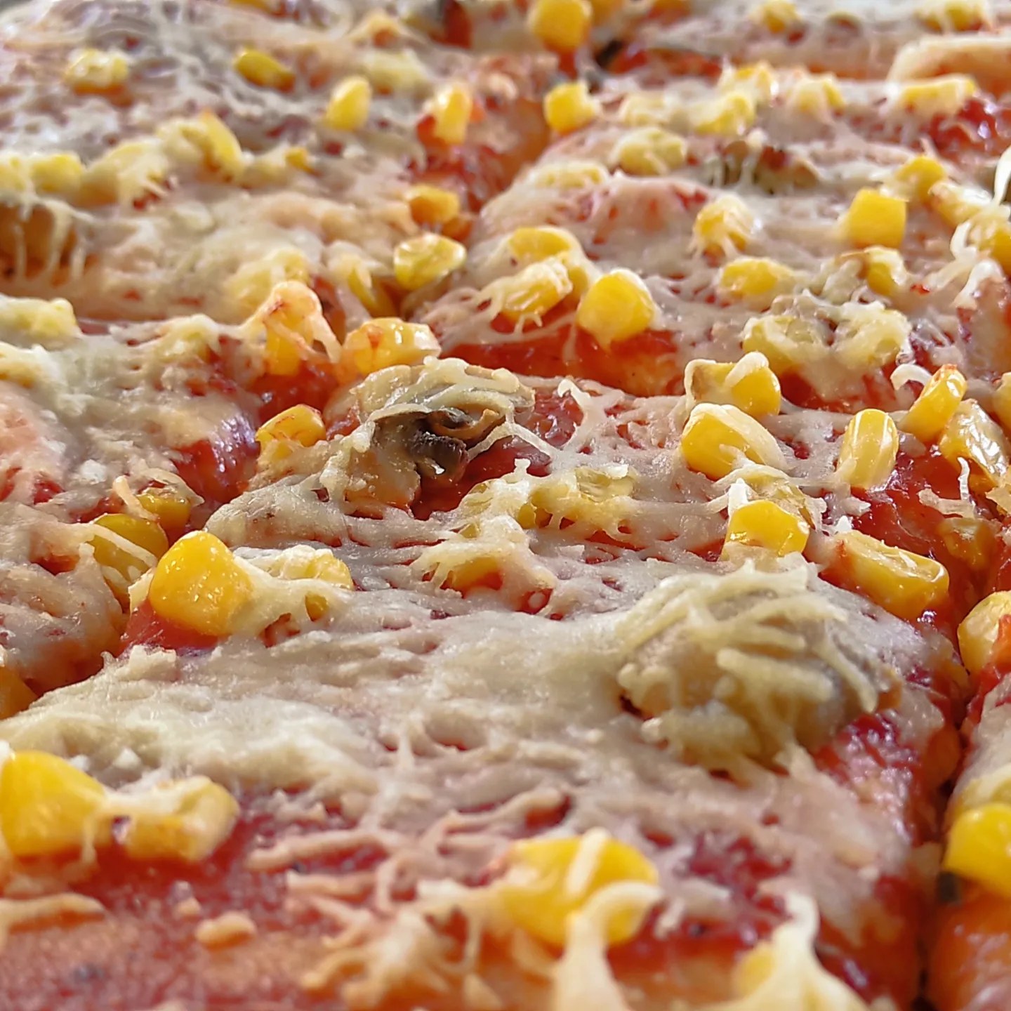Közeli kép egy pizzáról, tetején olvasztott sajttal, csemegekukoricával, gombával és paradicsomszósszal. A pizzát négyzet alakú darabokra vágják, a feltéteket egyenletesen elosztva a felületen, így részletes képet adnak az összetevőkről.
