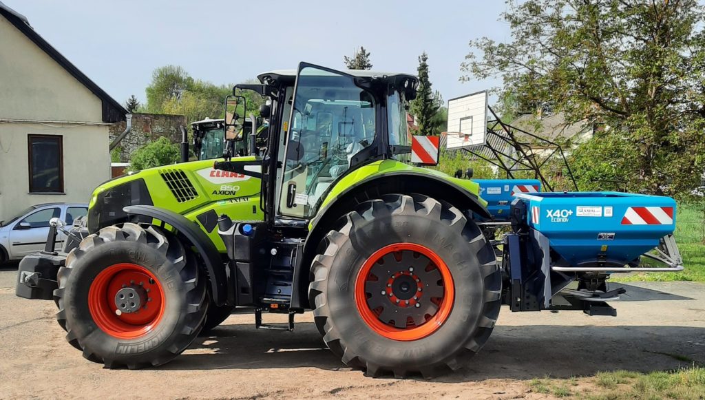 Egy zöld-fekete Claas Axion 850 traktor nagy narancssárga kerekekkel a szabadban parkol egy napsütéses napon. A hátuljára szerelt kék Kverneland X40 szórófejjel van felszerelve. A háttérben egy kosárlabda és zöld fák láthatók.