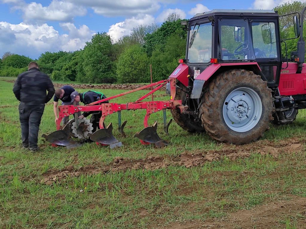 Két ember dolgozik egy piros traktoron, eke tartozékkal, zöld mezőn. Az egyik ember letérdel, hogy megvizsgálja az ekét, míg a másik a közelben áll. A traktor oldalra van elhelyezve, a háttérben fák és részben felhős az ég.