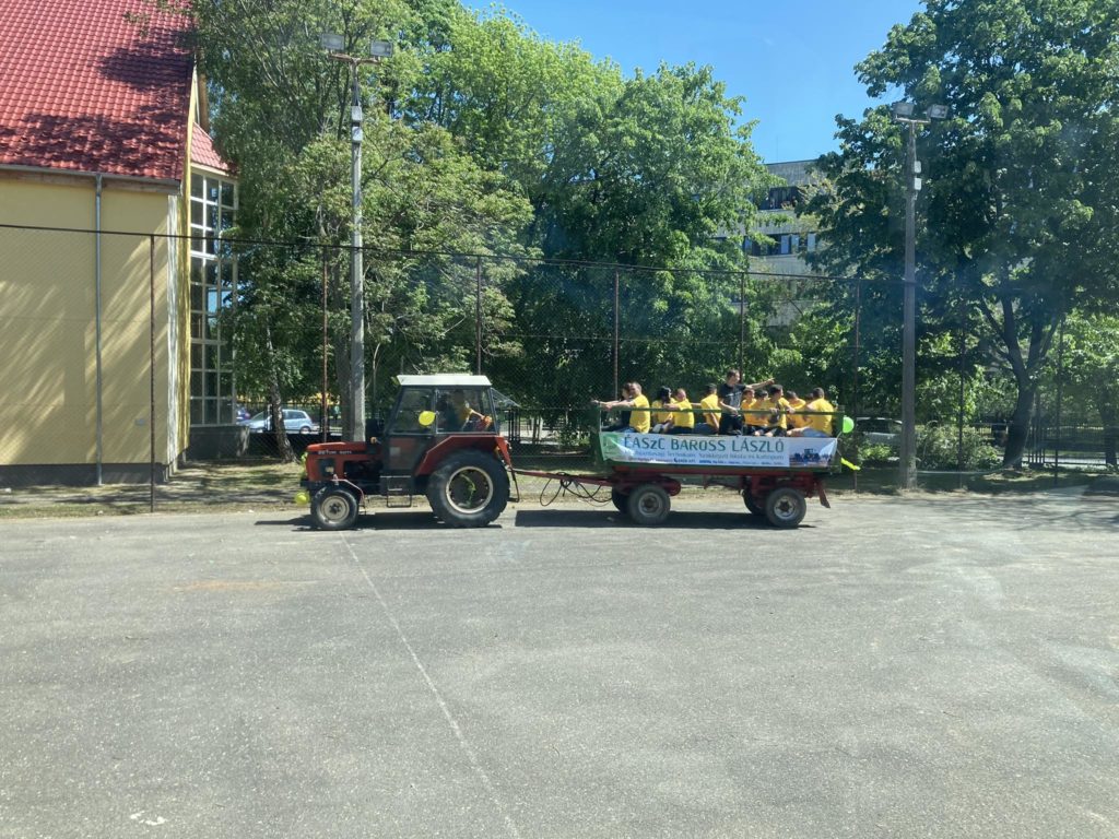 Egy piros traktor egy kis nyitott pótkocsit húz, tele sárga inget viselő emberekkel. Az előzetesen egy szalaghirdetés található, amely szöveges hirdetésnek vagy közleménynek tűnik. A háttérben fák és egy vörös tetős épület látható.