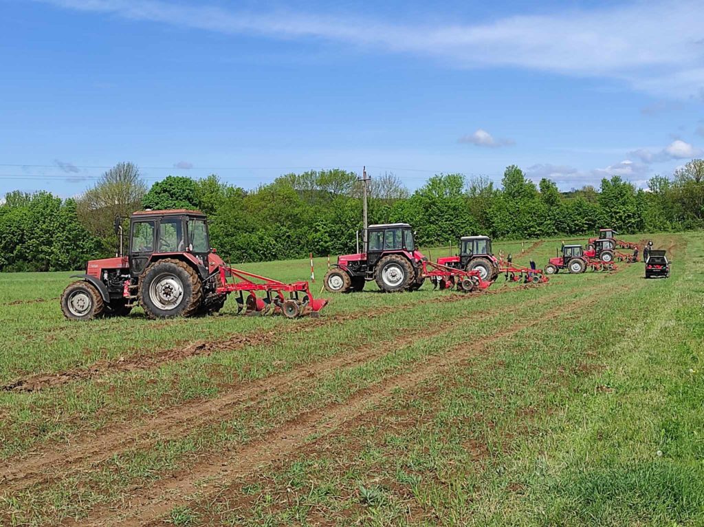 Négy piros traktor gazdálkodik egy zöld mezőn, mindegyiken egy-egy ember vesz részt egy másik traktor által húzott ekén, és lépcsőzetes sorban dolgozik. A mezőt sűrű zöld fák határolják a kék ég alatt, néhány elszórt felhővel.
