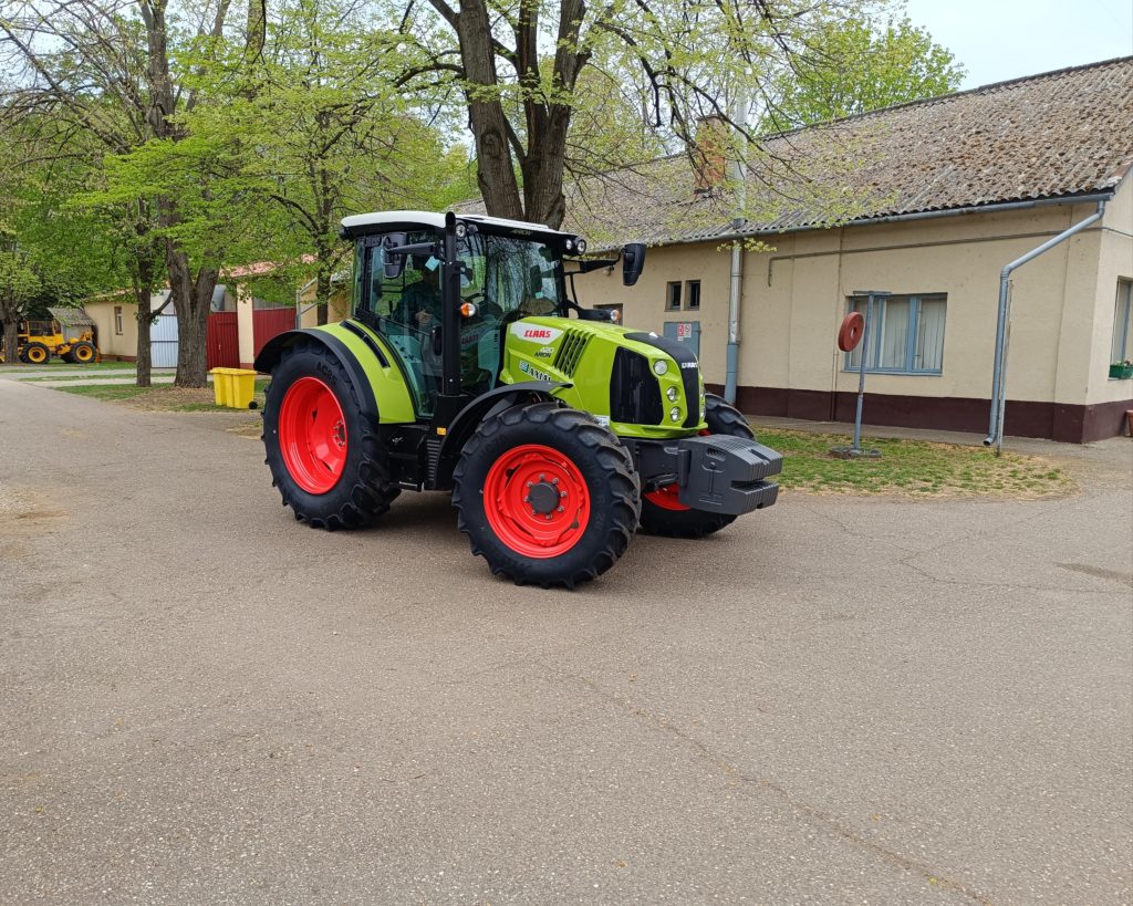 Egy aszfaltozott úton egy zöld-fekete traktor parkol nagy piros kerekekkel. A környéken található egy kopott tetős kis épület és néhány zöld lombozatú fa, ami azt jelzi, hogy tavasz van. Úgy tűnik, hogy a jelenet vidéki vagy tanyasi környezetben zajlik.