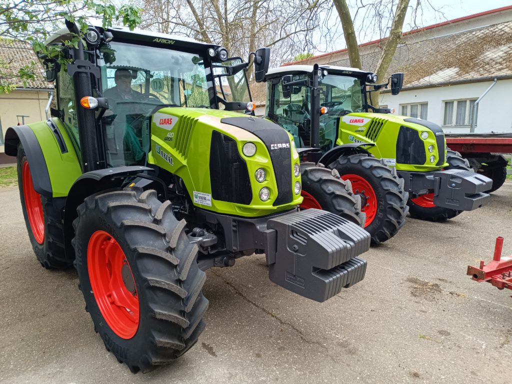Két zöld, élénkpiros kerekű Claas Arion 410 traktor parkolt betonfelületen. A háttérben fák és épületek láthatók. A traktorok nagy elülső súlyokkal és modern felszereltséggel vannak felszerelve, jelezve, hogy készen állnak a mezőgazdasági használatra.