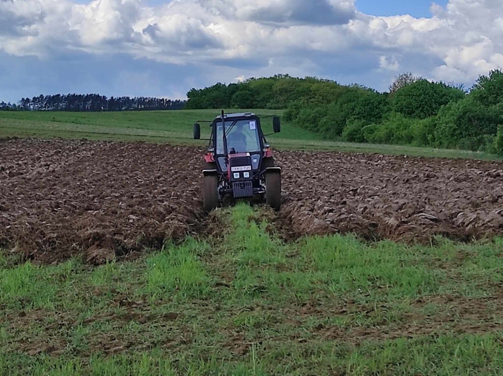 Egy traktor nagy szántóföldet szánt, kevert és megmunkált talajjal. Az ég részben felhős, a távolban fasor határolja a mezőt. A jelenet mezőgazdasági tevékenységet ábrázol vidéki környezetben.
