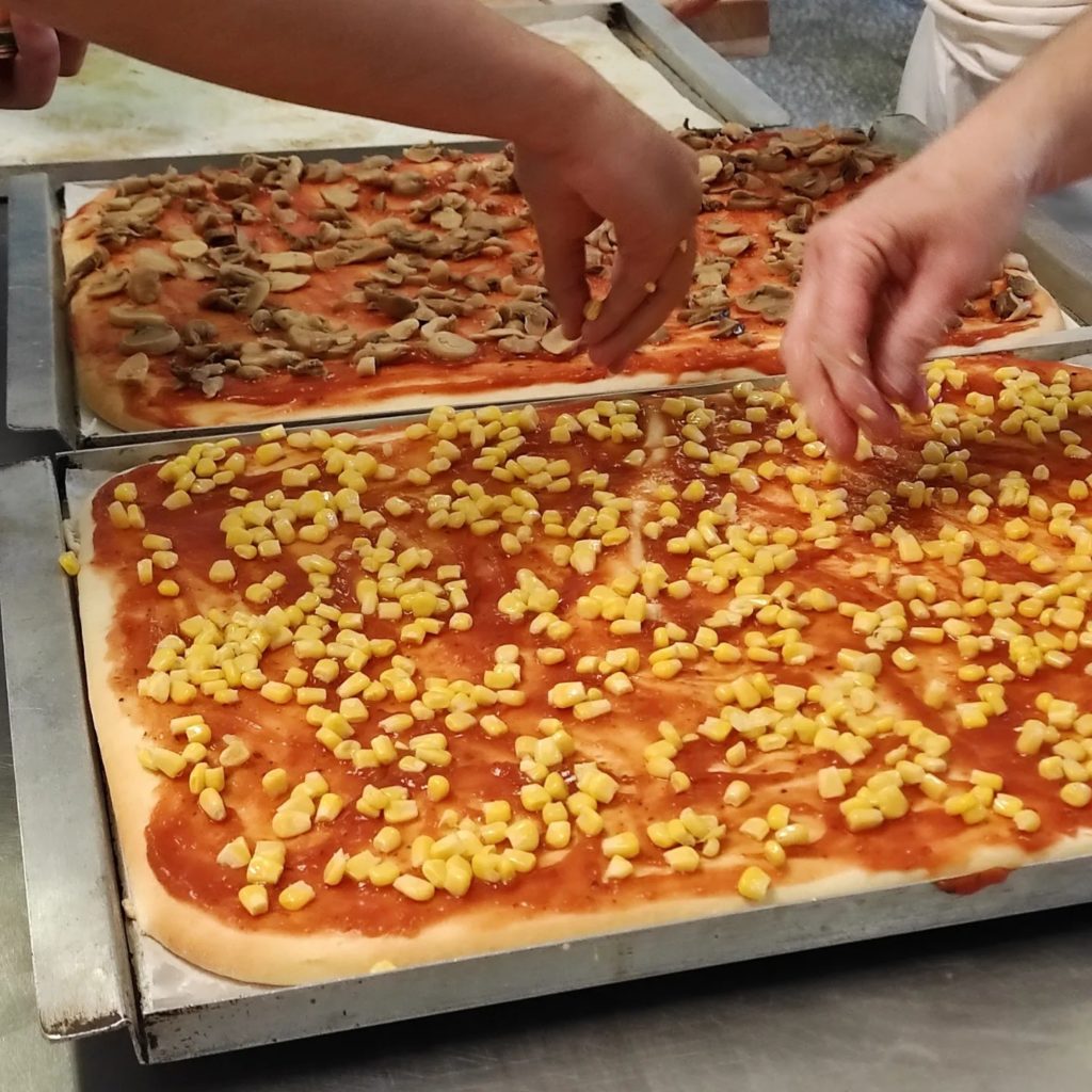Két téglalap alakú pizza készül. Az egyik pizza tetejére szeletelt gomba, a másikra kukoricaszem kerül. Két pár kéz teszi a feltétet a pizzákra konyhai környezetben.