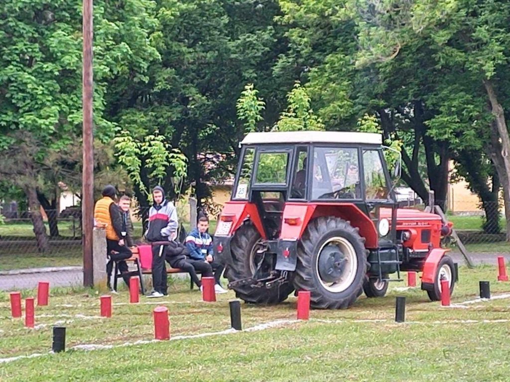 Egy csoport ember áll és ül egy füves területen parkoló piros traktor mellett. A traktor körül több függőleges farönk van elhelyezve. A háttérben fák és kerítés látható. Úgy tűnik, hogy a helyszín a szabadban van, esetleg egy parkban vagy egy vásártéren.