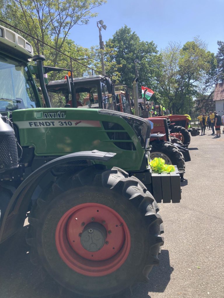 Színes traktorok sora, köztük egy zöld Fendt 310, amely egy napsütéses napon parkolt. A jelenet egy vidéki vagy mezőgazdasági területen játszódik, fákkal és emberekkel a háttérben. Az egyik traktoron zászló látható, amely egy kis ünnepi hangulatot ad.