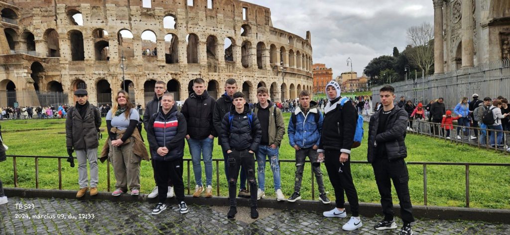 A római Colosseum előtt egy 11 fős csoport, többségében fiatal férfiak pózolnak egy fotóhoz. Alkalmi ruhát viselnek, és néhányan kabátot és kapucnis pulcsit viselnek. Felhősnek tűnik az idő, és más turisták is láthatók a háttérben.