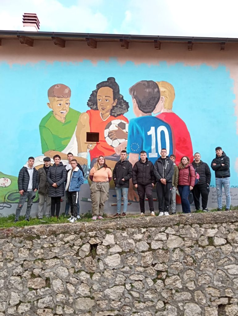 Egy embercsoport áll az épület falára festett falfestmény előtt. A falfestmény egy animált jelenetet ábrázol sportoló gyerekekről, az egyik gyerek focilabdát tart a kezében. A csoport egy kő emelvényen áll, és mosolyog a kamerába.