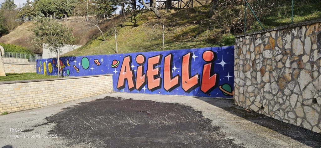Egy színes falfestmény egy kőfalon nagy, félkövér betűkkel az "Aielli" szót írja, kék háttér előtt csillagokkal és bolygókkal. A környező terület burkolt felületet, kőfalat és füves, fás lejtőt foglal magában.