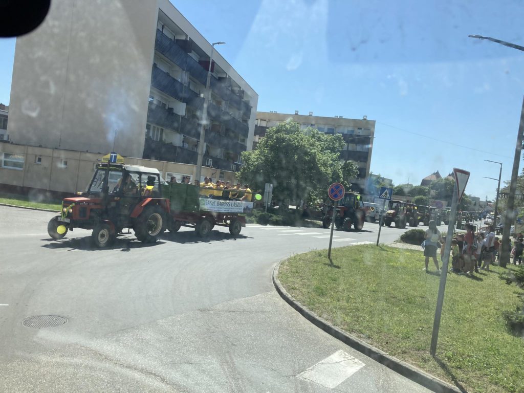 Traktorok felvonulása halad át egy városon, többen figyelik a járdát. A traktorok táblákat, díszeket hordoznak, lakóépületek sorakoznak az utcán. Az ég tiszta és napos.