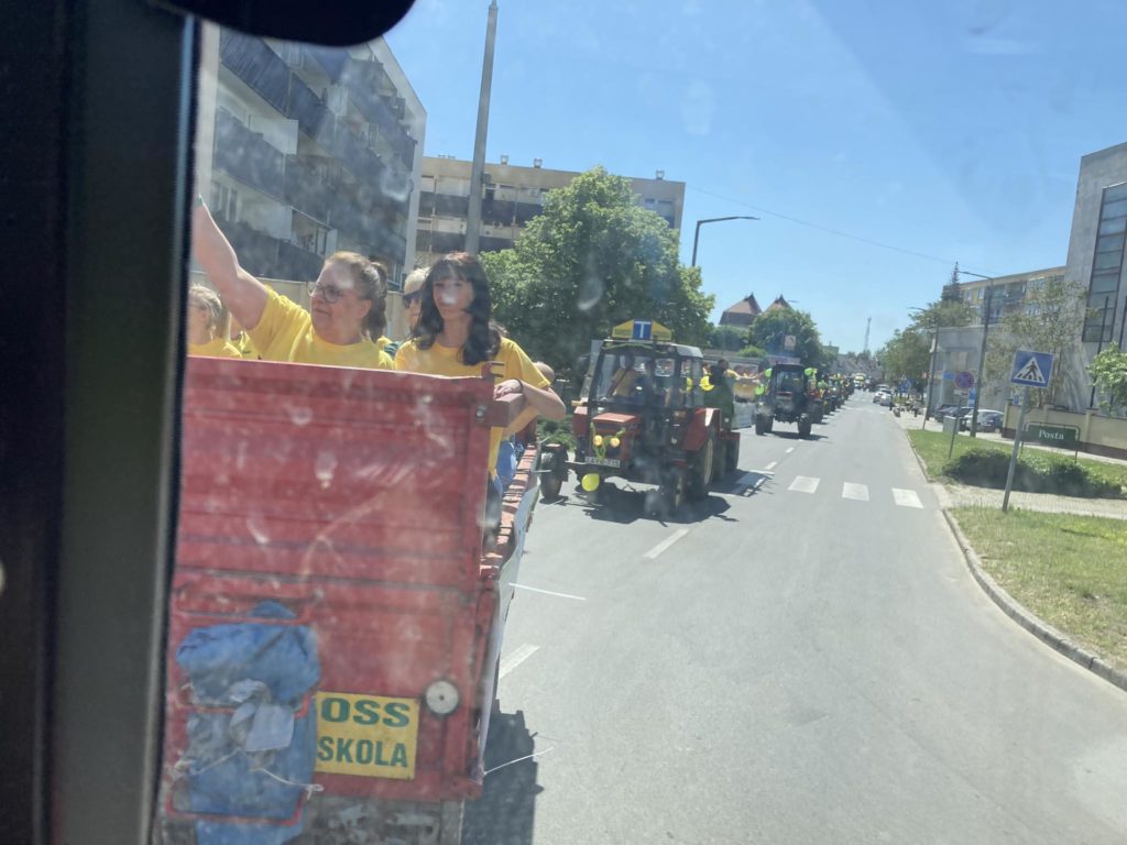 Sárga inget viselő emberek egy csoportja egy piros traktor hátán lovagol egy városi területen egy felvonulásnak vagy eseménynek tűnő utcán. Más traktorok követik őket. Épületek és fák szegélyezik az utcát, az emberek a járdákról figyelik.