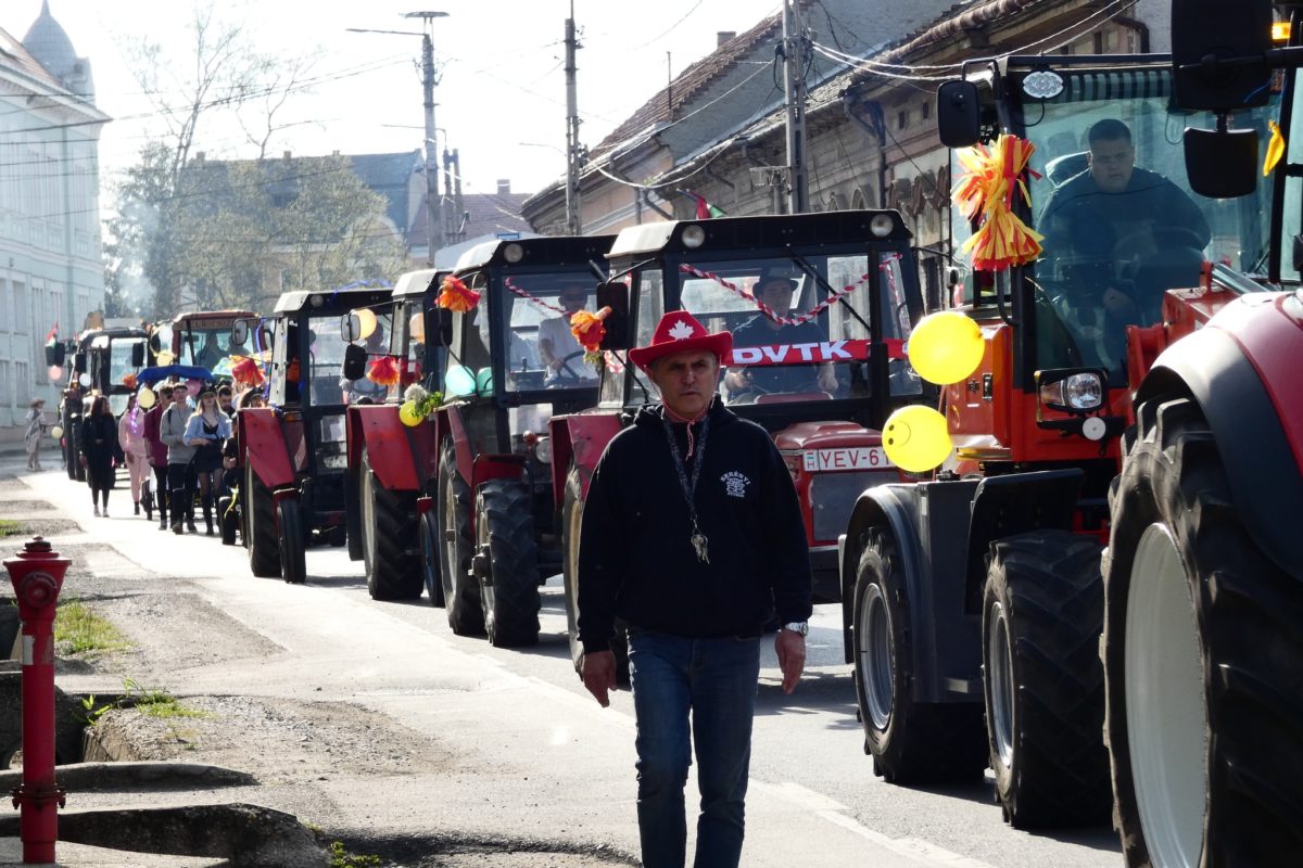 Egy férfi sétál az utcán a léggömbökkel és szalagokkal díszített traktorok sora előtt. Emberek láthatók a traktorokon ülni, a háttérben pedig házak sorakoznak az utcán. Úgy tűnik, ez egy helyi felvonulás vagy ünneplés.