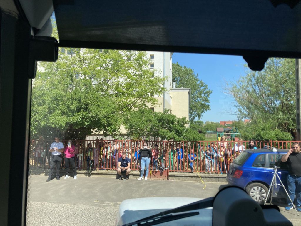 Kilátás egy jármű belsejéből, amely egy kerítés mögött álló emberek tömegét mutatja. Vannak, akik kamerát vagy felvevőkészüléket tartanak a kezükben. A háttérben fák és egy épület, jobb oldalon pedig egy parkoló kék autó látható.