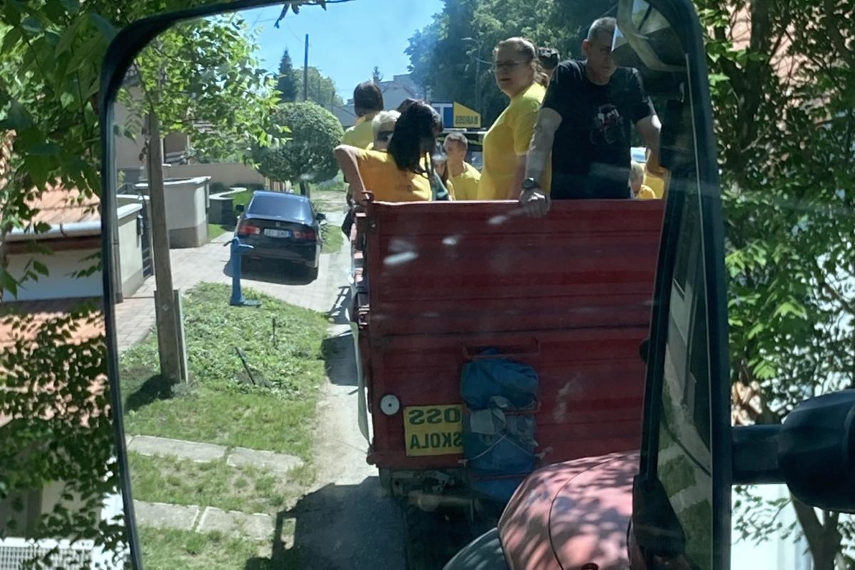 Sárga inget viselő emberek egy csoportja egy traktorra erősített piros pótkocsi hátuljában ül. A jelenet egy nagy oldaltükörben tükröződik. Egy ember látható az előtérben, aki fényképez, a háttérben pedig fák és házak.