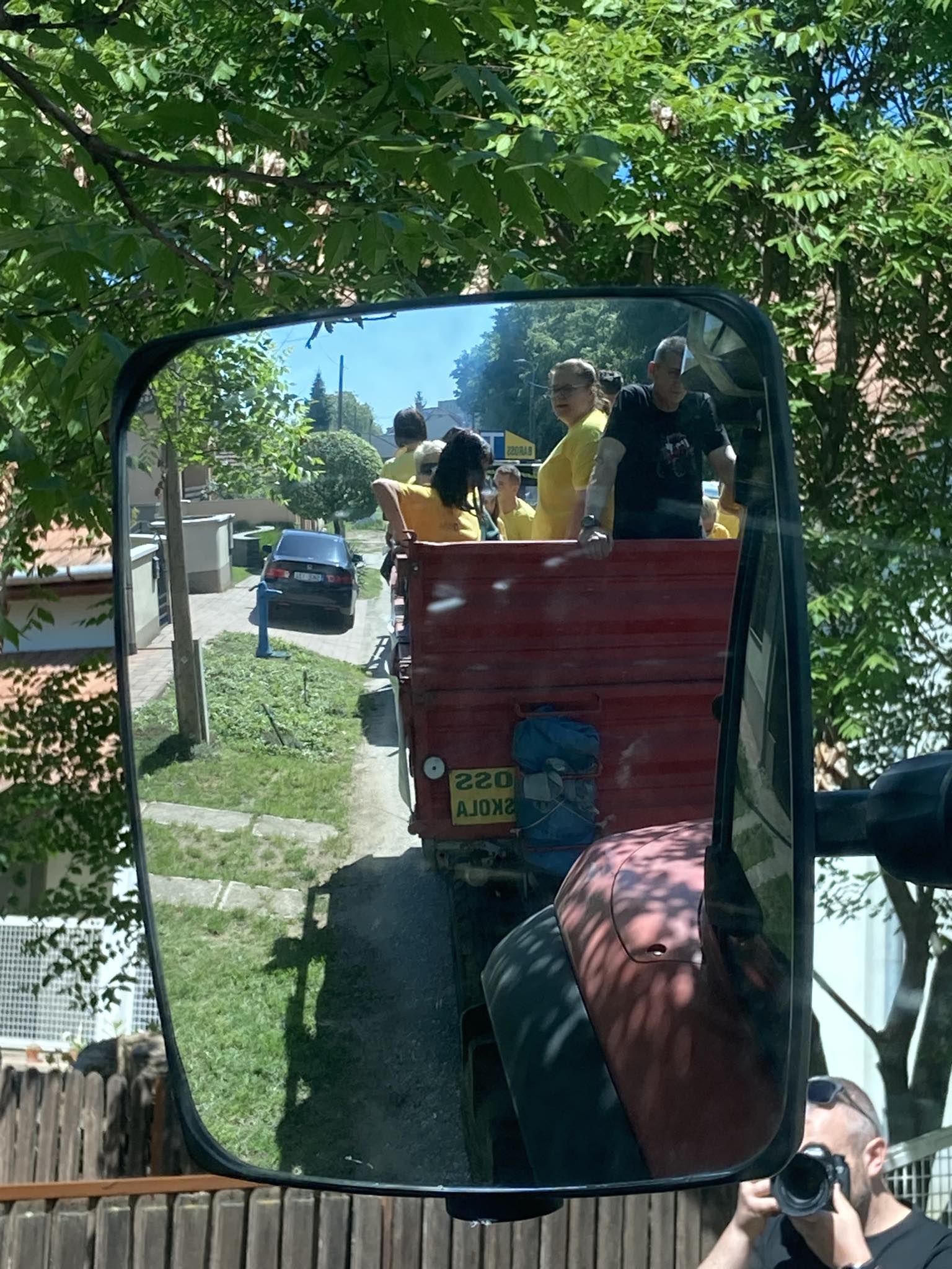 Sárga inget viselő emberek egy csoportja egy traktorra erősített piros pótkocsi hátuljában ül. A jelenet egy nagy oldaltükörben tükröződik. Egy ember látható az előtérben, aki fényképez, a háttérben pedig fák és házak.