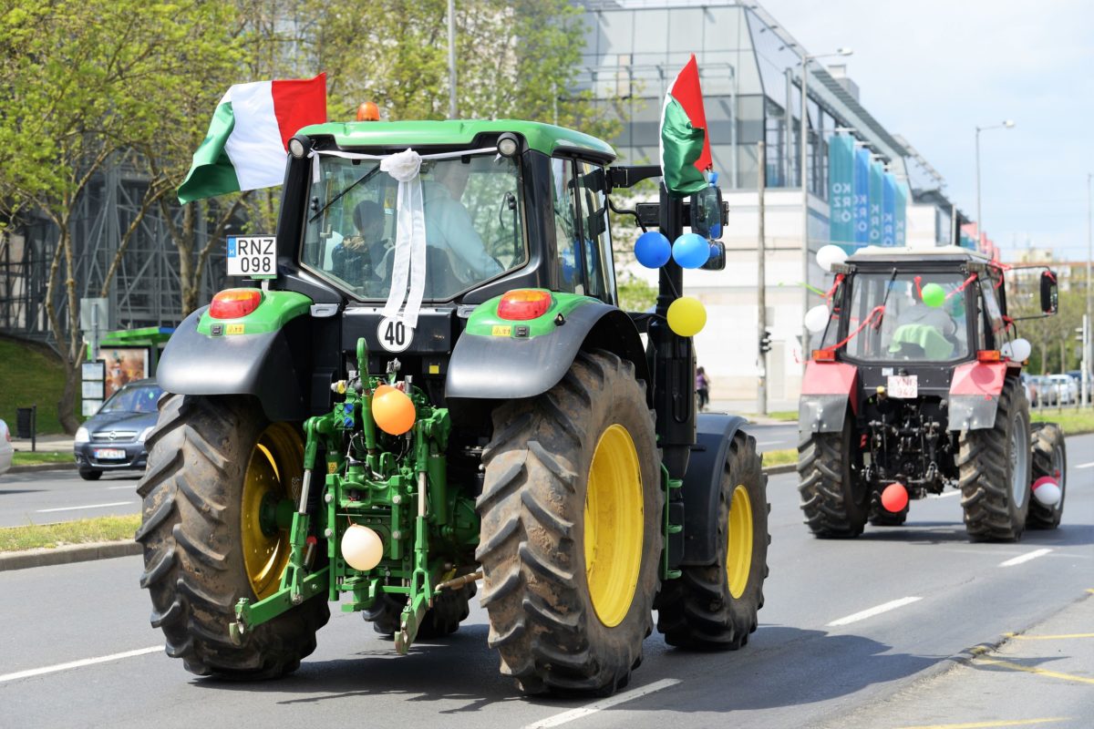 Két színes léggömbökkel és zászlókkal díszített traktor közlekedik a város egyik utcáján felvonulás közben. Az előtérben látható zöld traktor tetőjén jól látható zászlók láthatók, míg a piros traktor szorosan követi mögötte. A háttérben fák és modern épületek sorakoznak.