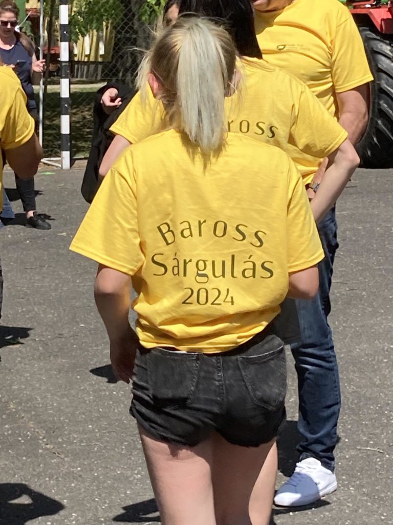 Egy szőke hajú, sárga pólót viselő, hátulján „Baross Sárgulás 2024” felirattal áll a szabadban. Más emberek veszik körül őket, akik hasonló inget viselnek. Az illető fekete rövidnadrágot is visel. A beállítás naposnak tűnik.