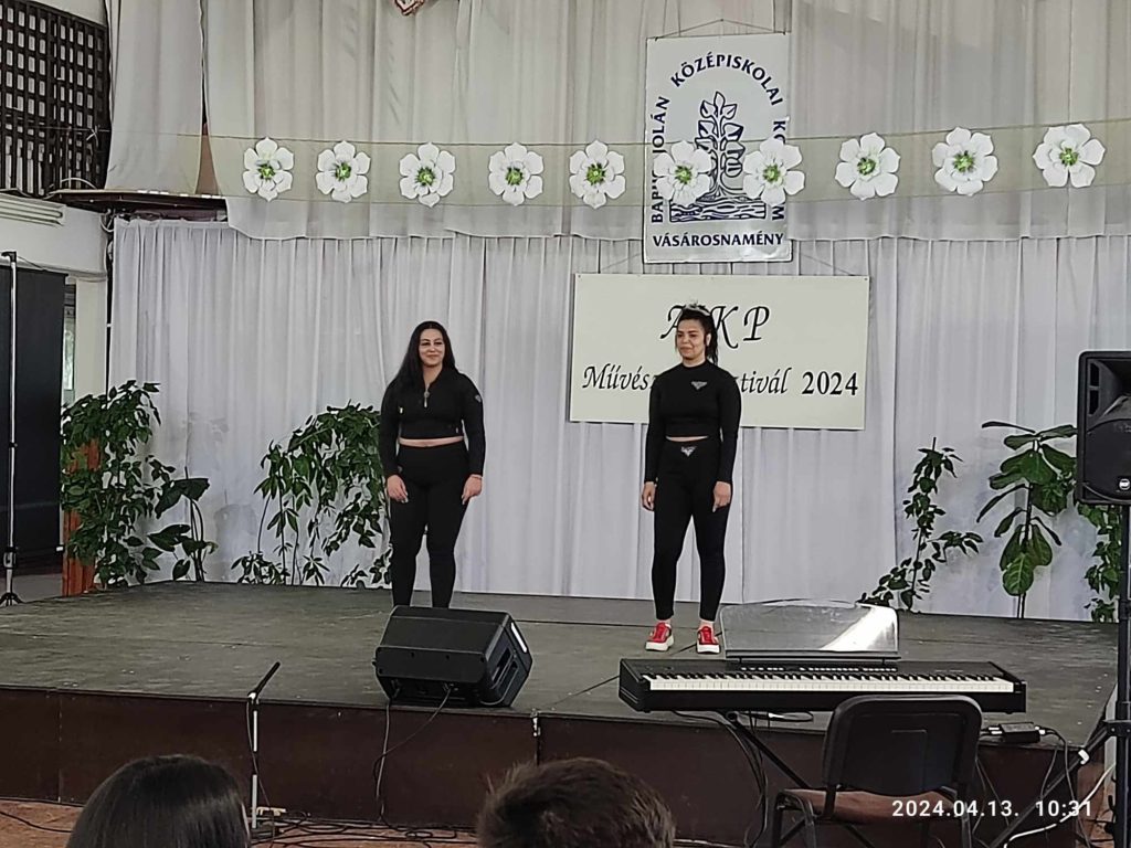 Két ember áll a virágokkal, transzparenssel és növényekkel díszített színpadon. Fekete ruhát viselnek, és szembefordulnak a közönséggel. A zenei berendezések, köztük a billentyűzet és a hangszórók láthatók. A szalaghirdetésen szöveg és dátum szerepel: 2024. április 13.
