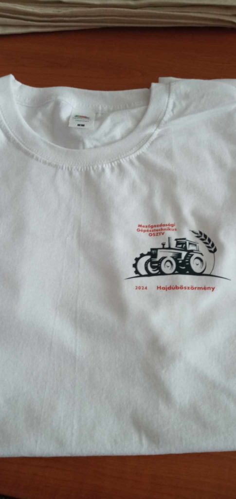 Fehér póló, a bal mellkason mintával. A terv egy traktor grafikáját, a "Mezőgazdasági Gépkiállítás 2014 Hajdúböszörmény" feliratot pirossal, a szöveg felett pedig egy kis logót tartalmazza. A pólót fa felületre helyezzük.