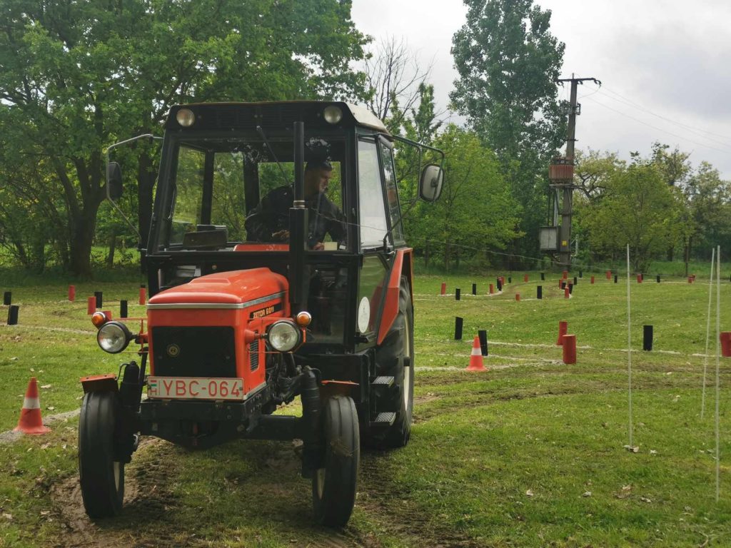 Egy piros-fekete traktor, benne sofőrrel közlekedik a különféle kúpokkal és oszlopokkal jelzett füves területen. A háttérben a felhős ég alatt fák és közműoszlop látható. A traktor rendszámtábláján ez áll: „YBC-064.