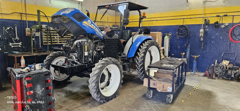 Egy szerelő egy nyitott motorháztetővel rendelkező kék traktoron dolgozik egy műhelyben. A műhelyben falakon lógnak a szerszámok, körülötte különféle felszerelések és dobozok. A szerelő arccal a traktor motorjára koncentrál.