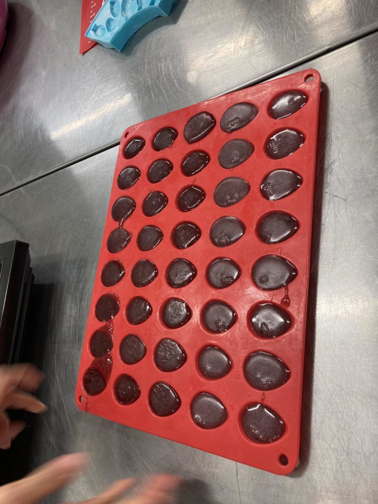 Sötét, ovális formájú csokoládét tartalmazó piros szilikon forma fémes felületen helyezkedik el. Egy személy keze részben látható a kép bal alsó sarkában, ami arra utal, hogy valószínűleg a csokoládéval dolgozik.