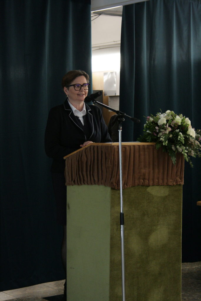 Egy nő áll az emelvényen mikrofonnal, és beszédet mond. Szemüveget, fekete öltönyt és fehér blúzt visel. A pódiumot barna kendő és virágkompozíció díszíti. A háttérben sötétzöld függönyök lógnak.