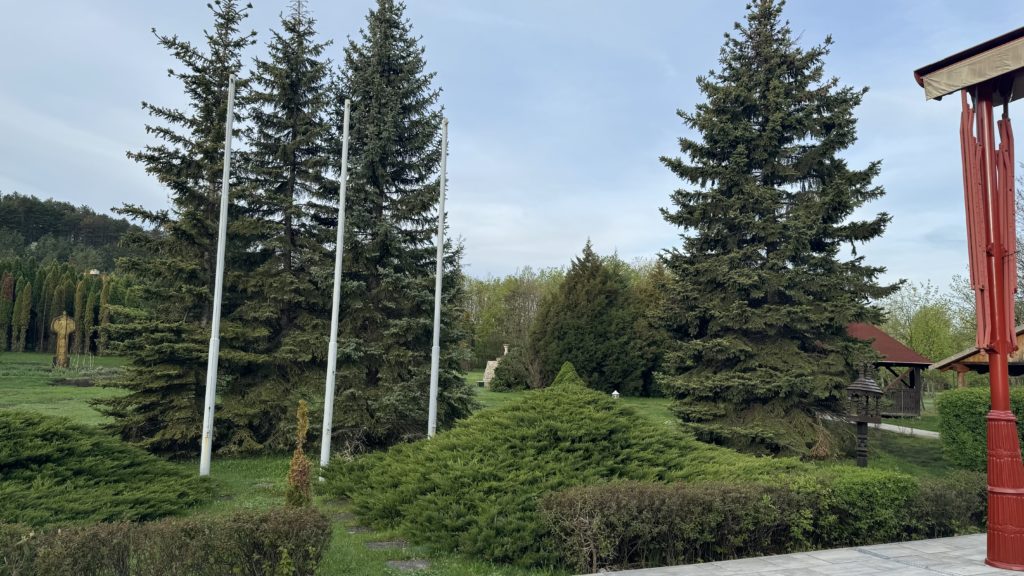 Buja zöld park számos magas örökzöld fával és három üres zászlórúddal. A tágas terület cserjésekkel és kőúttal parkosított. Jobb oldalon piros építmény látható, részben felhős az ég.