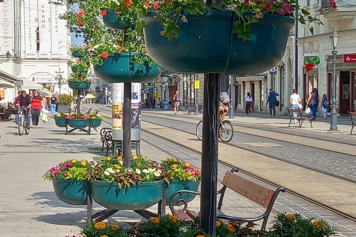 Színes virágokkal teli többszintes virágtartókkal szegélyezett városi utca. Az utca világosszürke téglával burkolt, és villamosvágányok futnak át rajta. A háttérben padok és sétáló-bicikliző emberek állnak a napsütéses égbolt alatt.