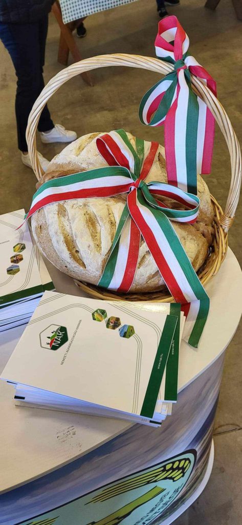 Az asztalon egy fonott kosár, amely egy nagy vekni kenyeret tartalmaz, piros, fehér és zöld szalaggal díszítve. A közelben található néhány zöld-fehér borítójú brosúra logókkal és szöveggel. Emberek állnak a háttérben.