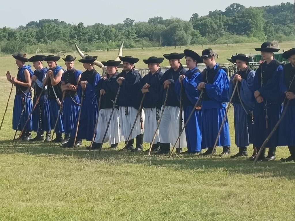 Hagyományos kék-fekete magyar pásztorruhába öltözött embercsoport áll sorban, hosszú fabotot tartva. Kint vannak egy füves mezőn, a háttérben fákkal.