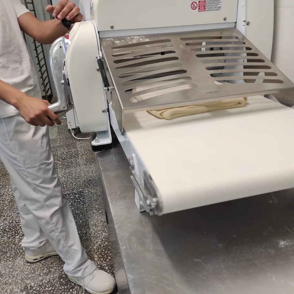 Egy fehér ruhás ember tésztalapítót üzemeltet a konyhában. Kézi hajtókart forgatnak, miközben vékony tésztarétegek haladnak át a gépen egy szállítószalagra. Úgy tűnik, hogy a helyszín egy kereskedelmi vagy ipari konyha.