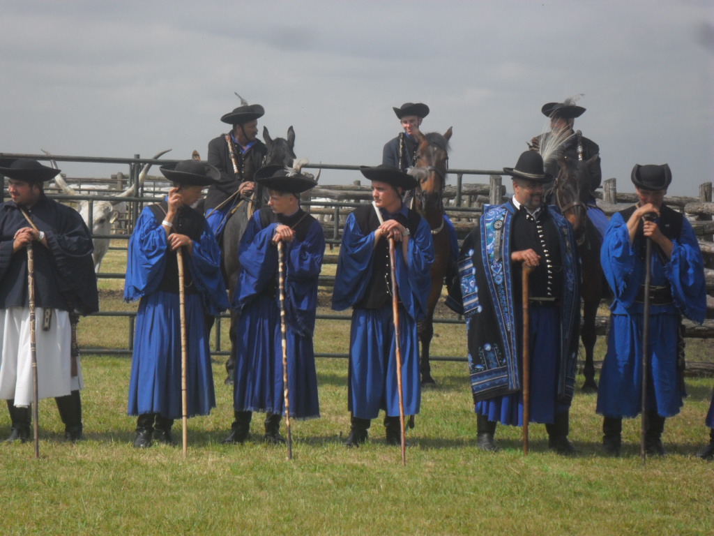 Hagyományos kék jelmezbe és fekete kalapba öltözött férfiak egy csoportja áll a sorban, hosszú fabotokkal. Mögöttük szintén hagyományos öltözetben lovas férfiak. A háttérben fából készült kerítés és enyhén borús ég látható.