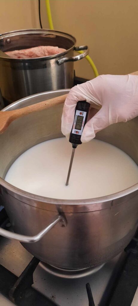 Kesztyűs kéz digitális ételhőmérőt tart egy főzőlapon lévő tejes fazékban, a kijelzőn 87,6 Celsius fokos hőmérséklet látható. Egy másik edény étellel van a háttérben.