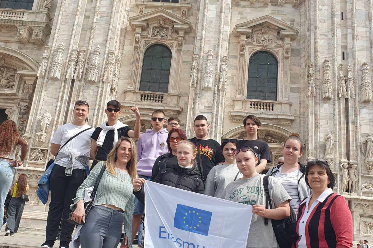 Emberek egy csoportja áll egy nagy, díszes katedrális előtt, kezükben az „Erasmus+” feliratú zászlóval az EU emblémájával. A katedrális bonyolult építészeti részletekkel, nagy boltíves ablakokkal és tornyos tornyokkal rendelkezik. Néhány csoporttag pózol, míg mások lazán mosolyognak.