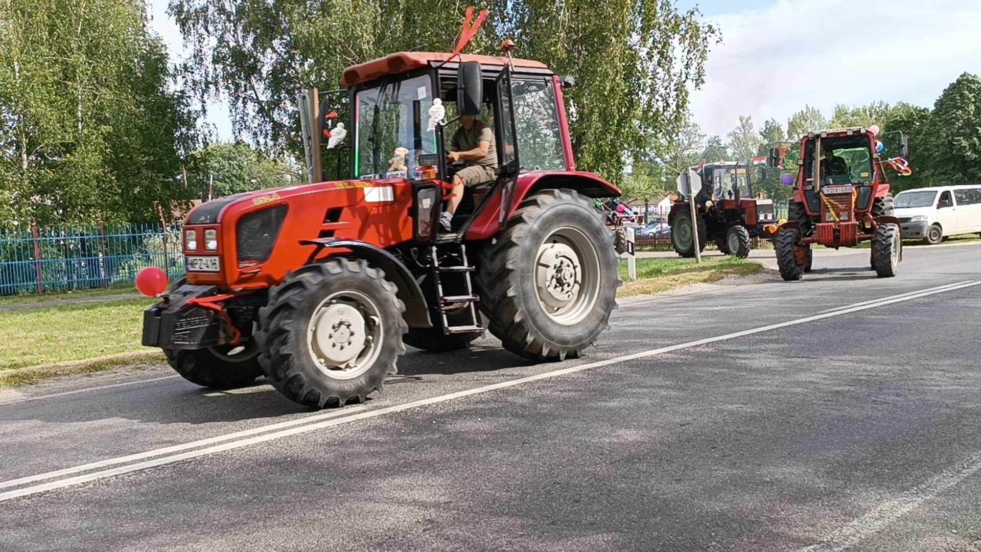 Egy piros traktor sofőrrel halad egy aszfaltozott úton felvonulás vagy esemény közben. Egy másik hasonló traktor következik mögötte. Az út széléről néznek emberek, a háttérben fák és kerítés látható.