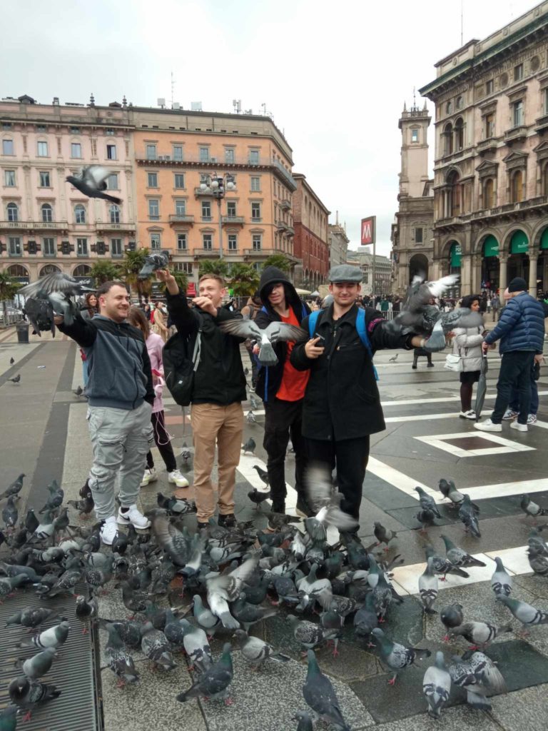 Egy csoport ember áll egy nyüzsgő városi téren, galambokkal körülvéve. Az előtérben négy személy mosolyog, és a madarakkal foglalkozik, a háttérben történelmi épületek és járókelők láthatók. A jelenet élénknek és vidámnak tűnik.