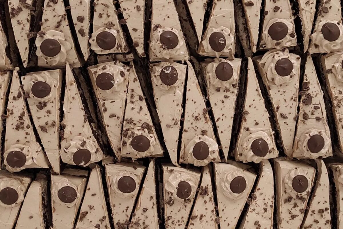 Szépen elrendezett csokoládétorták, világos színű cukormázzal és egy-egy kerek csokoládédarabbal minden szeleten. A sütemények szorosan egymás mellett helyezkednek el, függőleges mintában, vonzó és rendezett megjelenést kölcsönözve.