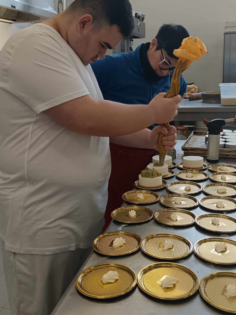 Két személy egy konyhában desszerteket készít. A bal oldali ember pipazacskót tart, és több tálcán elhelyezett kis tésztalapokra tejszínt nyom. A jobb oldali személy szemüveget és kék inget visel, és figyeli a folyamatot.