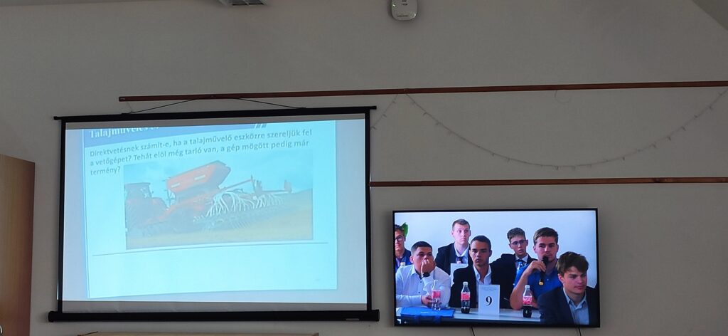 A bal oldali vetítővászon mezőgazdasági gépek képe látható magyar szöveggel. A jobb oldalon a televízió képernyőjén fehér inges férfiak és fiúk csoportja látható, akik egy asztalnál ülnek italokkal és egy 9-es számmal, és figyelmesen néznek valamit.
