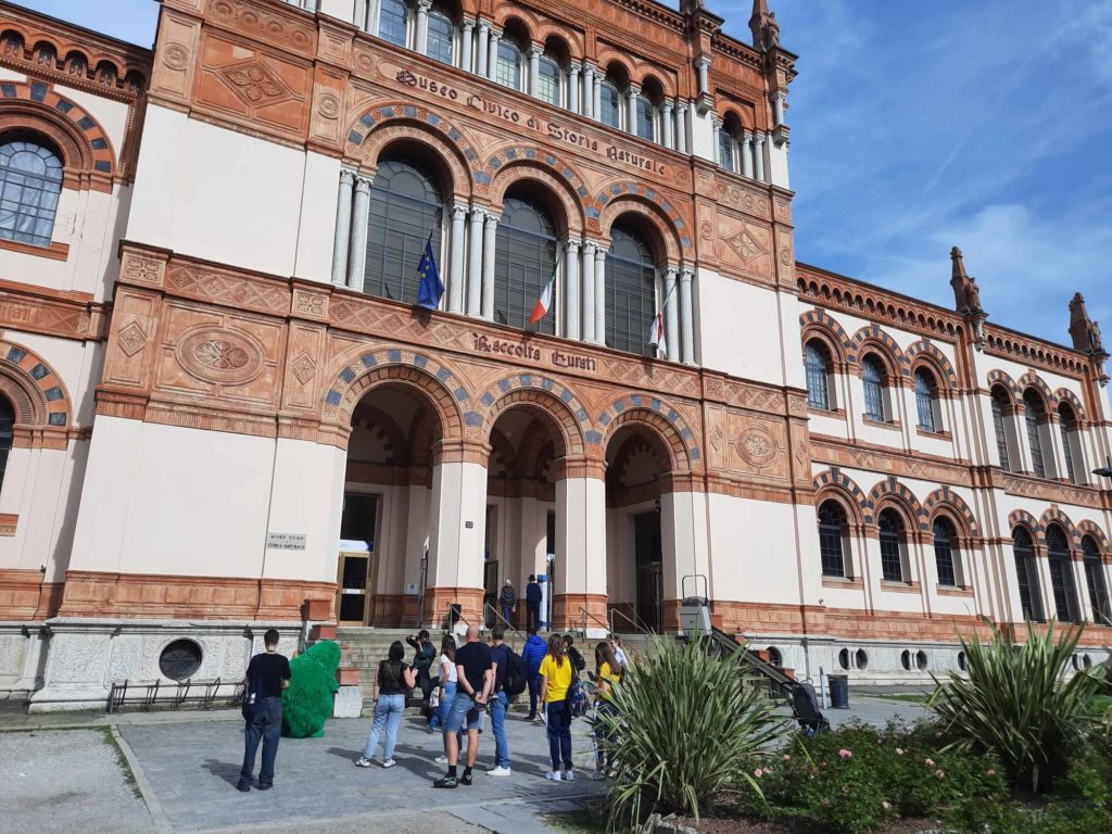 Emberek egy csoportja egy nagy, díszes építészetű történelmi épület bejáratánál. Az épület boltíves ablakai, dekoratív téglafalazat, a bejárat fölött a "Scuola Media Statale" felirat látható. A háttérben a kék ég látható.